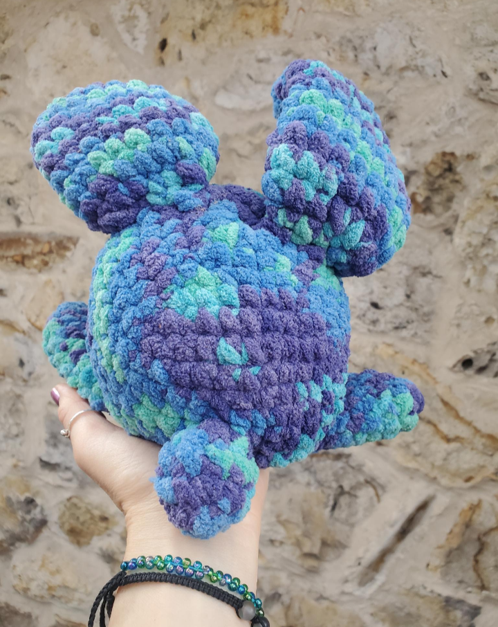 Hand holding a blue and green crocheted plush bunny toy against a stone wall background