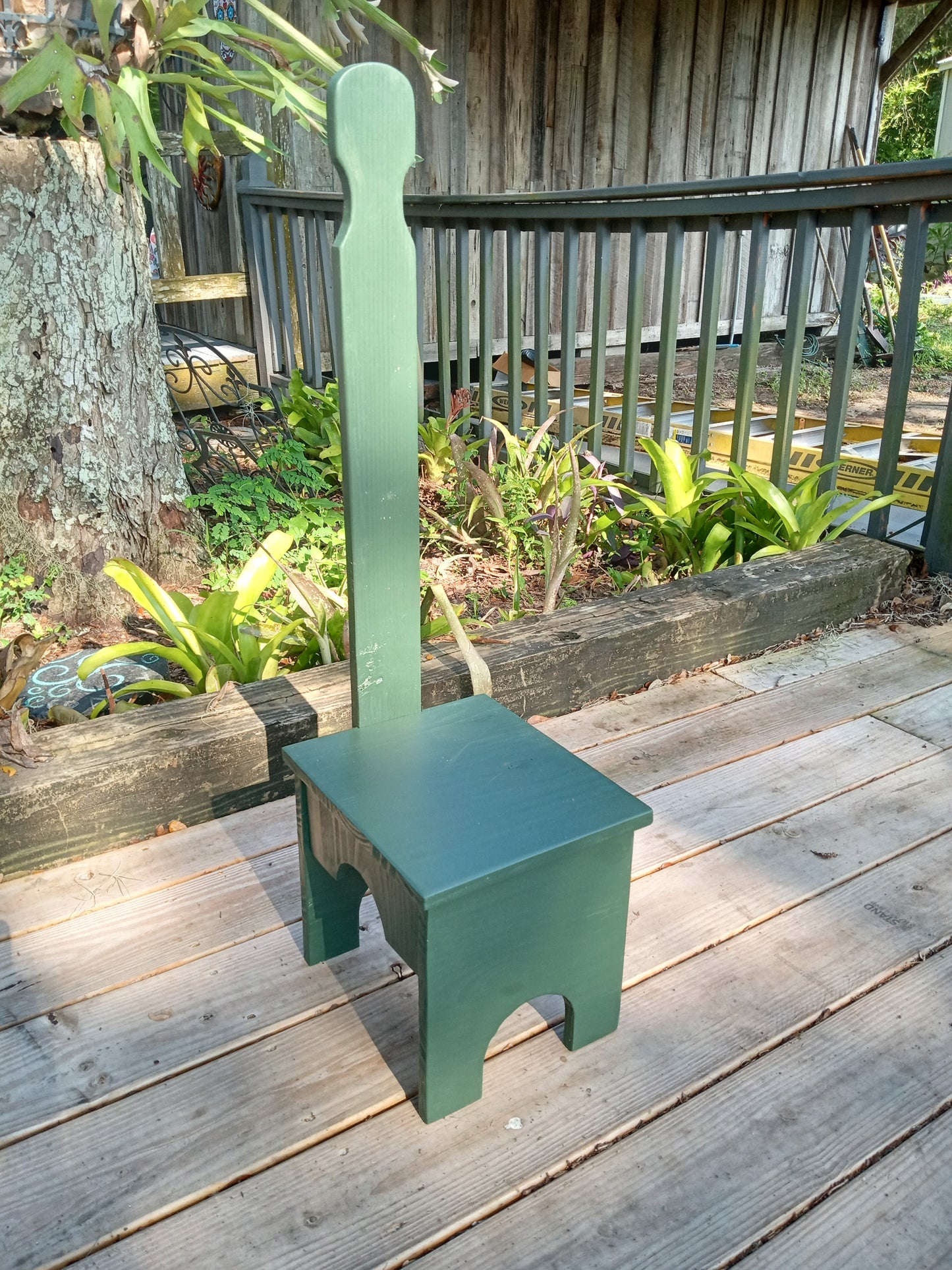 One stable step stool painted green and standing in an outdoor setting with a garden in the background.