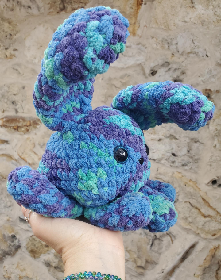 Handmade blue and green crochet bunny toy held against a textured beige wall.