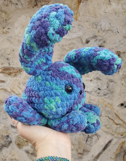 Handmade blue and green crochet bunny toy held against a textured beige wall.