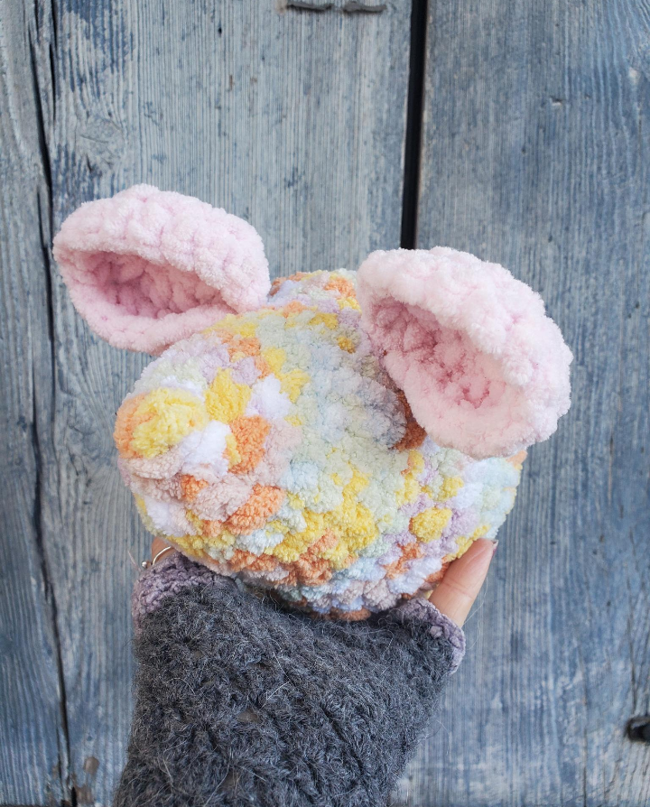 Hand holding a colorful, fluffy crochet mouse head against a wooden background
