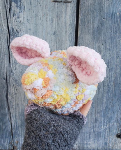 Hand holding a colorful, fluffy crochet mouse head against a wooden background