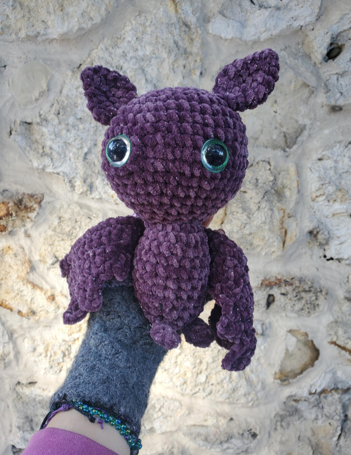 Purple crochet bat toy held by a gloved hand against a stone wall background