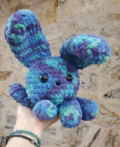 Crocheted blue and purple bunny held against a stone wall background