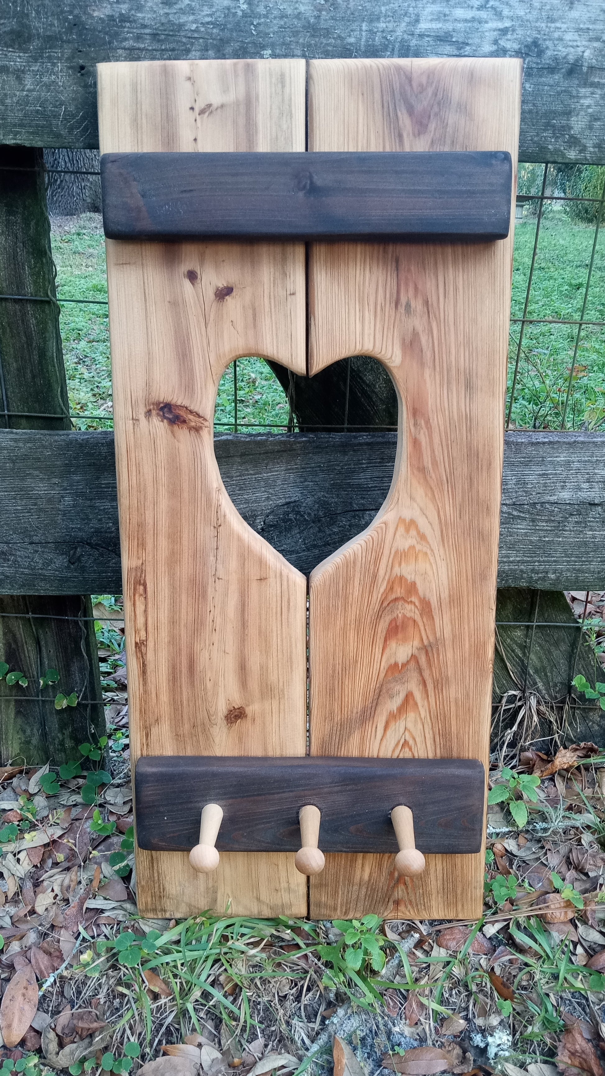 Cypress Wall Decor Coat Rack with heart cutout, featuring three wooden pegs and rustic panel design, leaning against a fence.