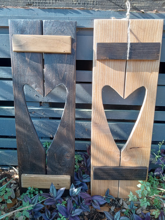 Two wooden decorative heart shapes with a cut-out in the center, one stained dark brown and the other in a light natural wood color, standing upright against a wooden fence with plants in the background.