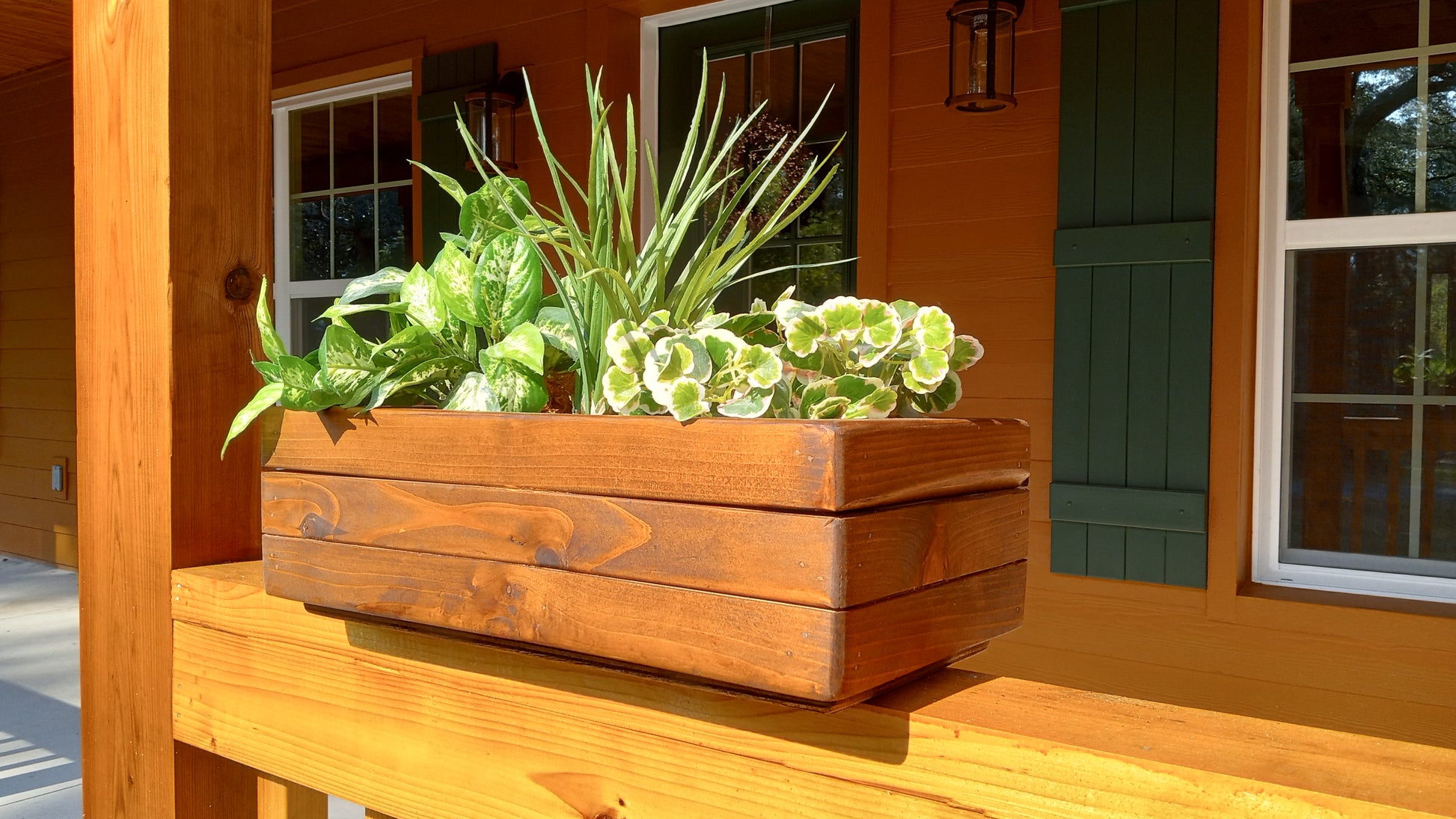 Large Cedar Planter Box on a porch railing, filled with varied green foliage, showcasing handcrafted cedar construction with mitered corners for strength.