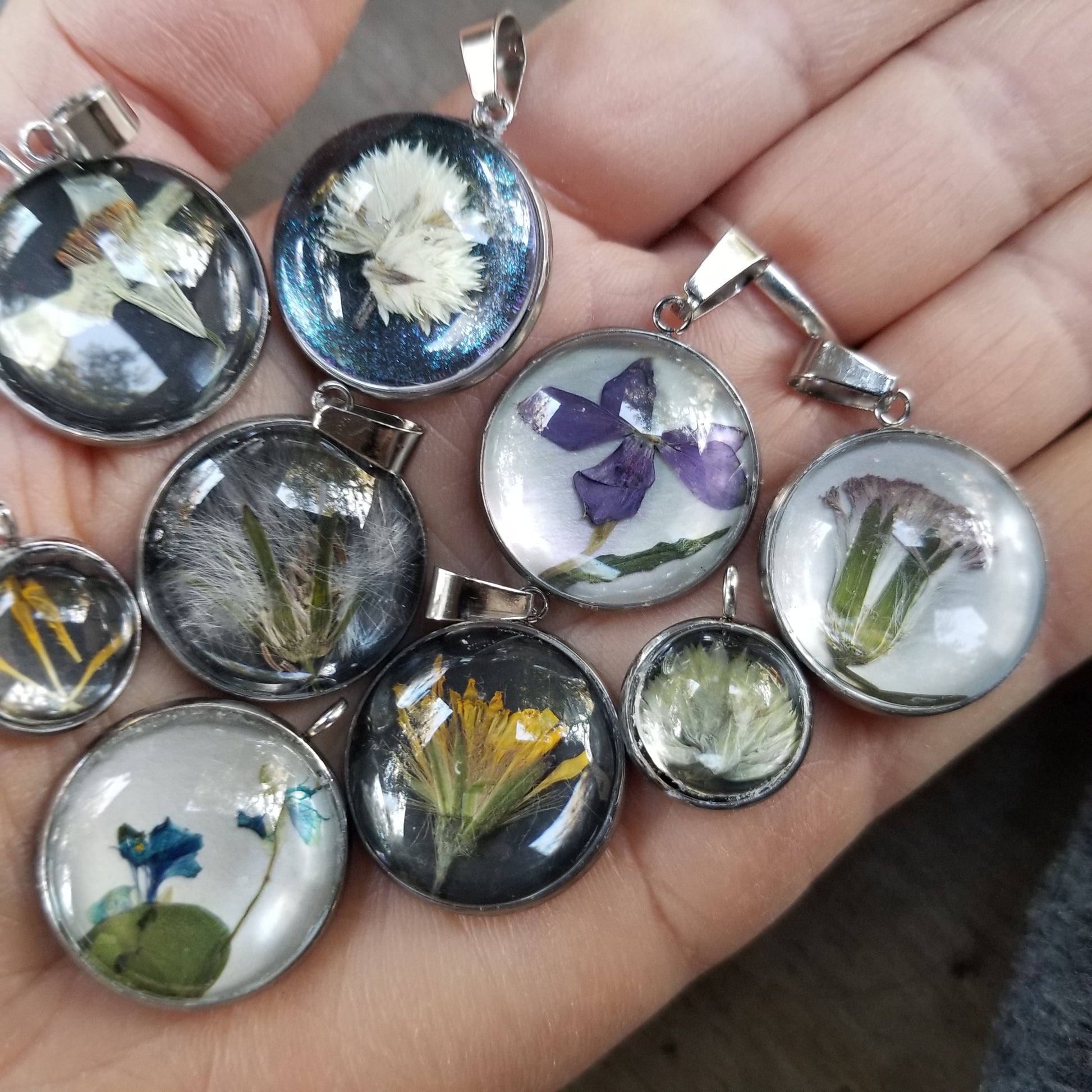 A hand holding several round pressed flower pendants with different flowers visible inside the pendants.