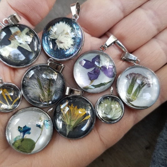 A hand holding several round pressed flower pendants with different flowers visible inside the pendants.