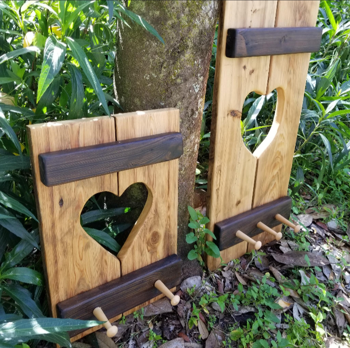 Cypress Wall Decor Coat Rack featuring two rustic wooden panels with heart cutouts, three pegs, and horizontal accents, leaning against a tree outdoors.