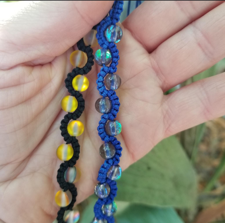 Two handmade waterfall bracelets held in a person's hand, one with black cord and yellow beads, and the other with blue cord and multicolored beads.